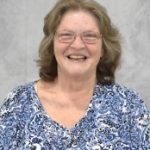 Woman in floral shirt, light brown, shoulder length hair with neutral gray background.