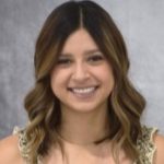 Woman with light-brown hair, wearing a floral top, sitting in front of a neutral, gray background.